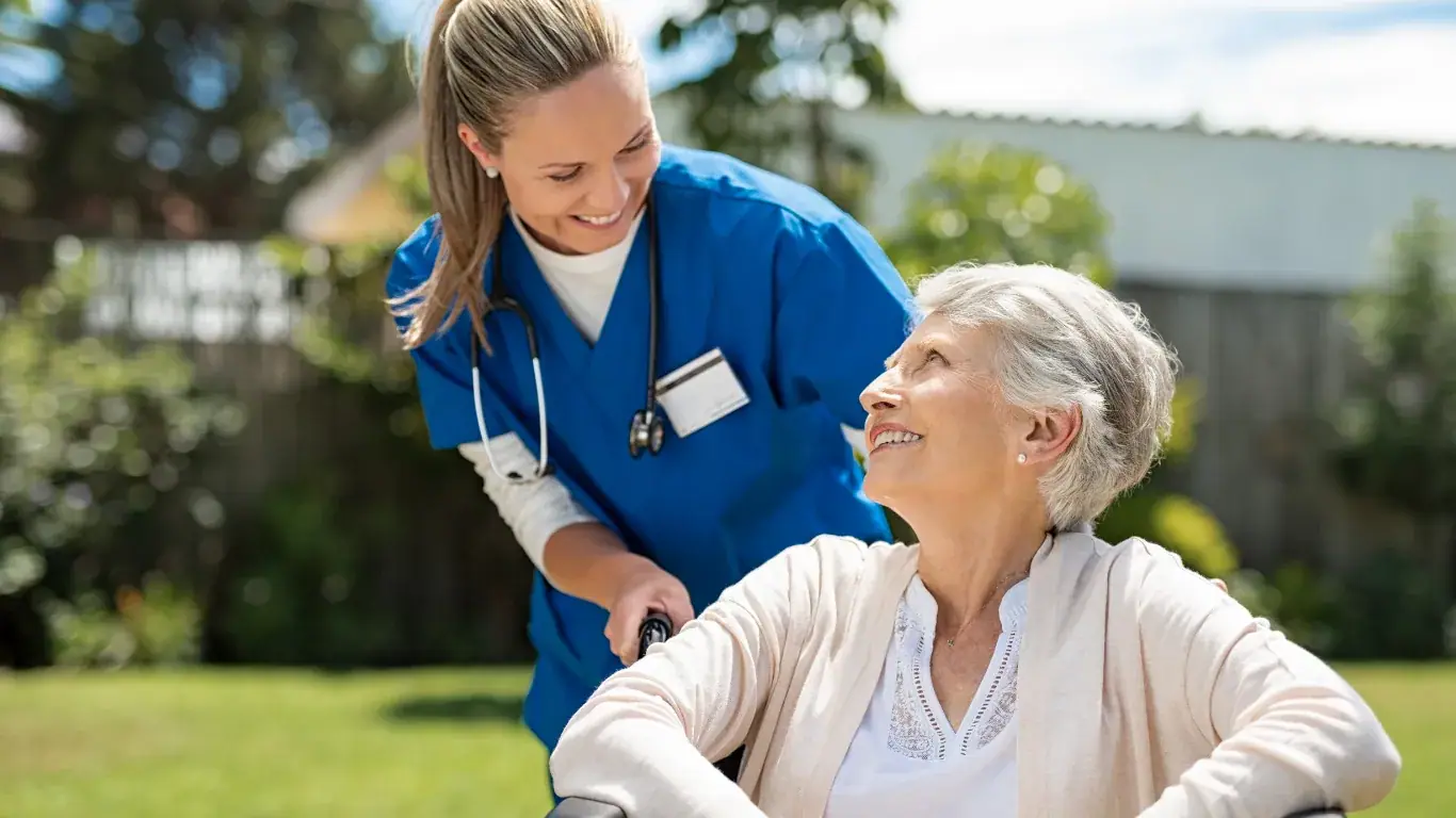 A carer in blue scrubs smiling with an elderly woman in a wheelchair outdoors